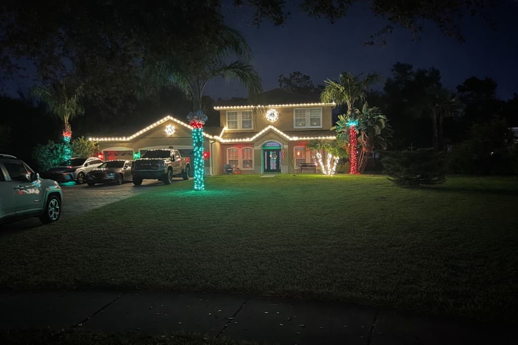 Two story home with soft white roofline lighting and tall palms wrapped in green and red lights
